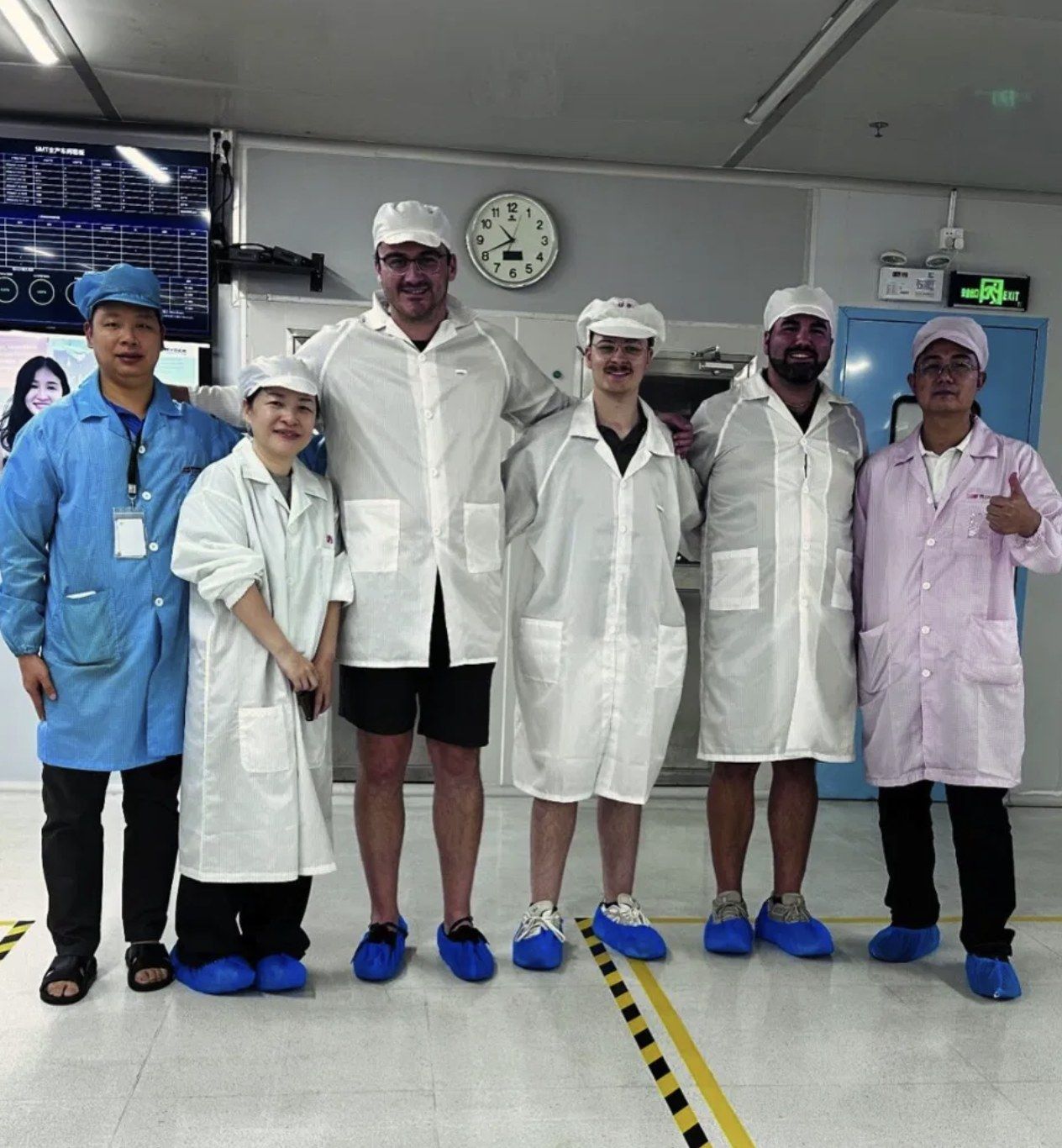 Six people in clean-room coats and shoe covers on a circuit board assembly floor.
