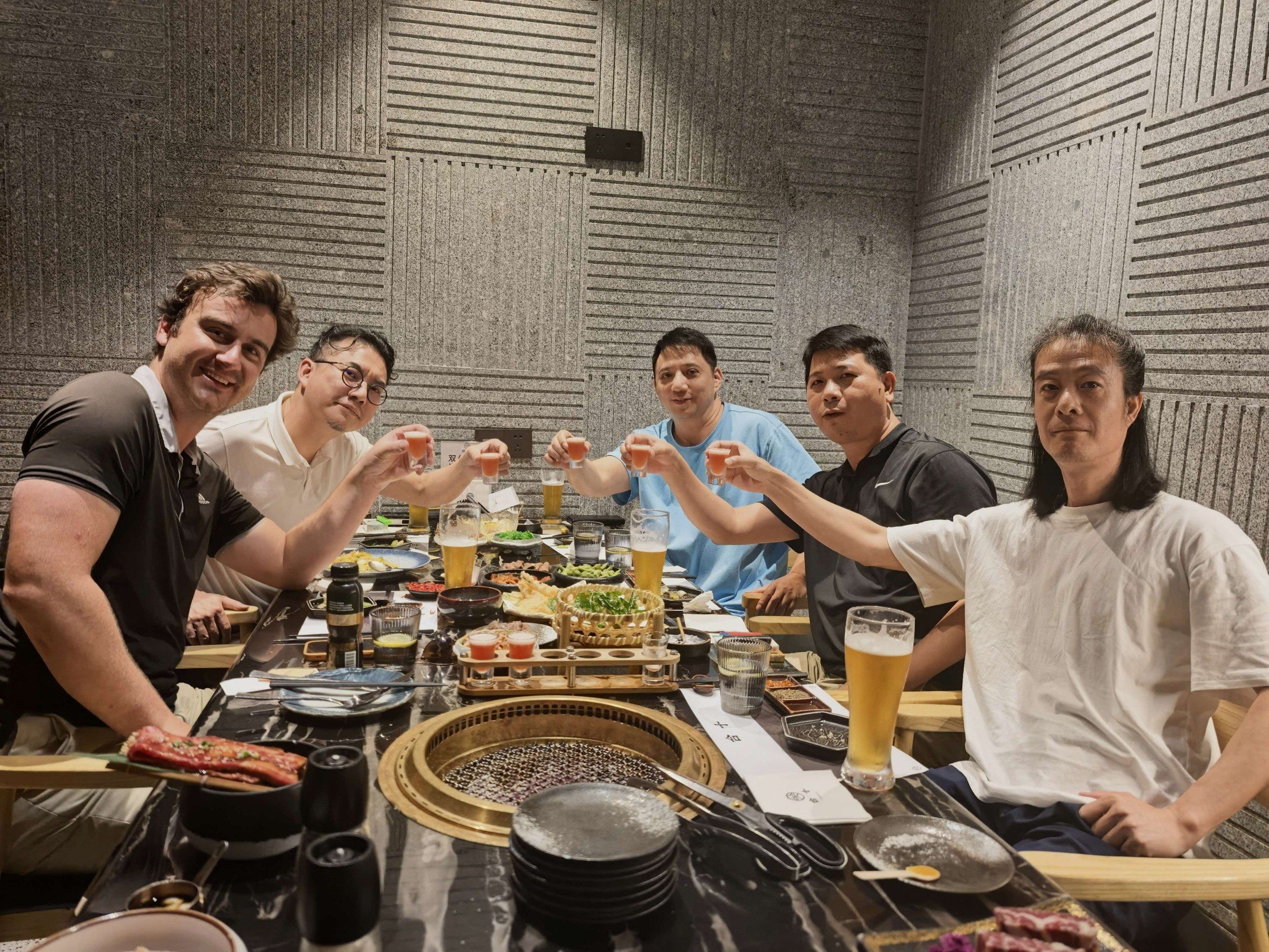 Curtis and four factory partners raising glasses at a barbecue table.