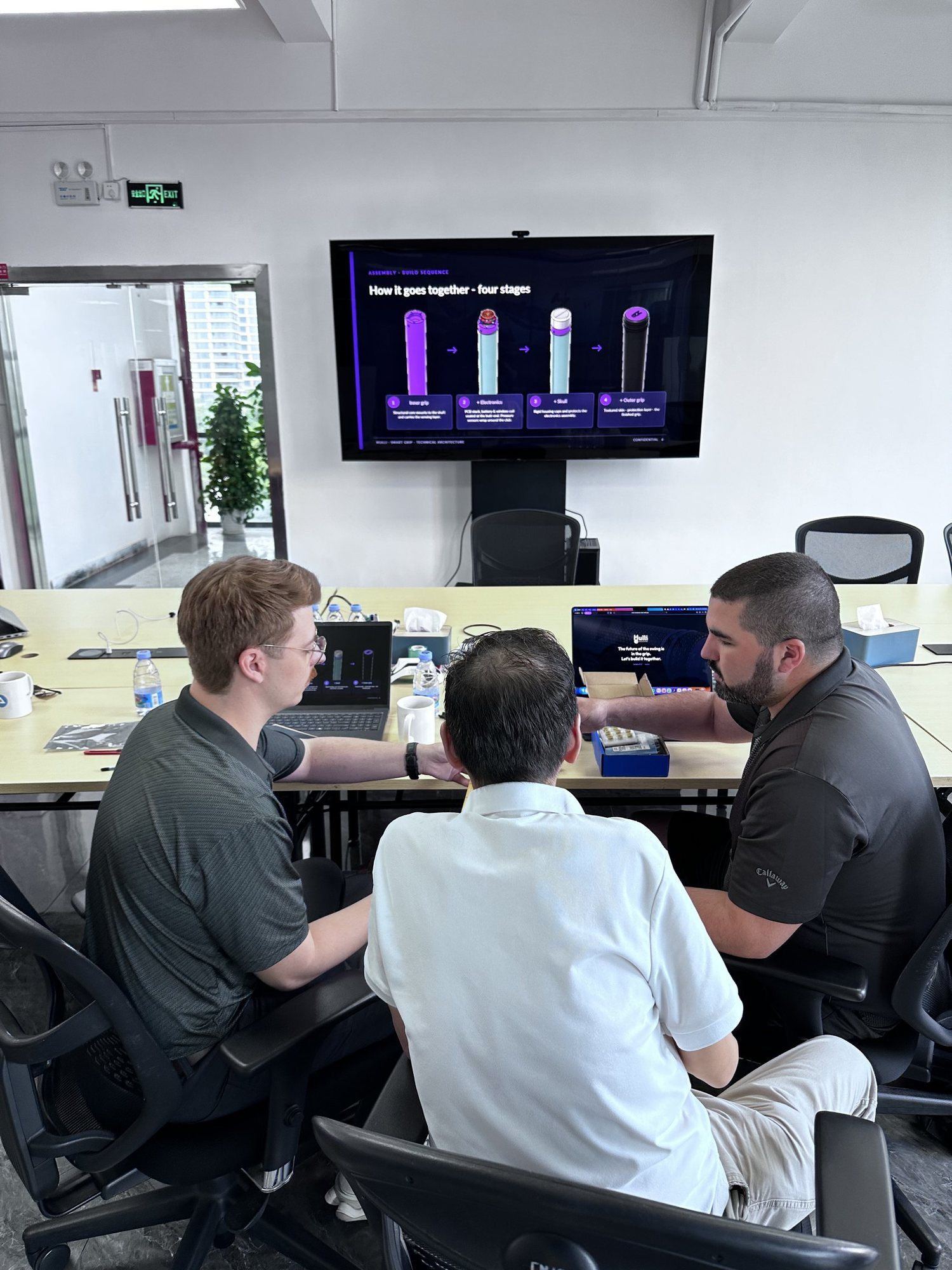 Founders across a meeting table from a factory manager, with a presentation on the screen behind.