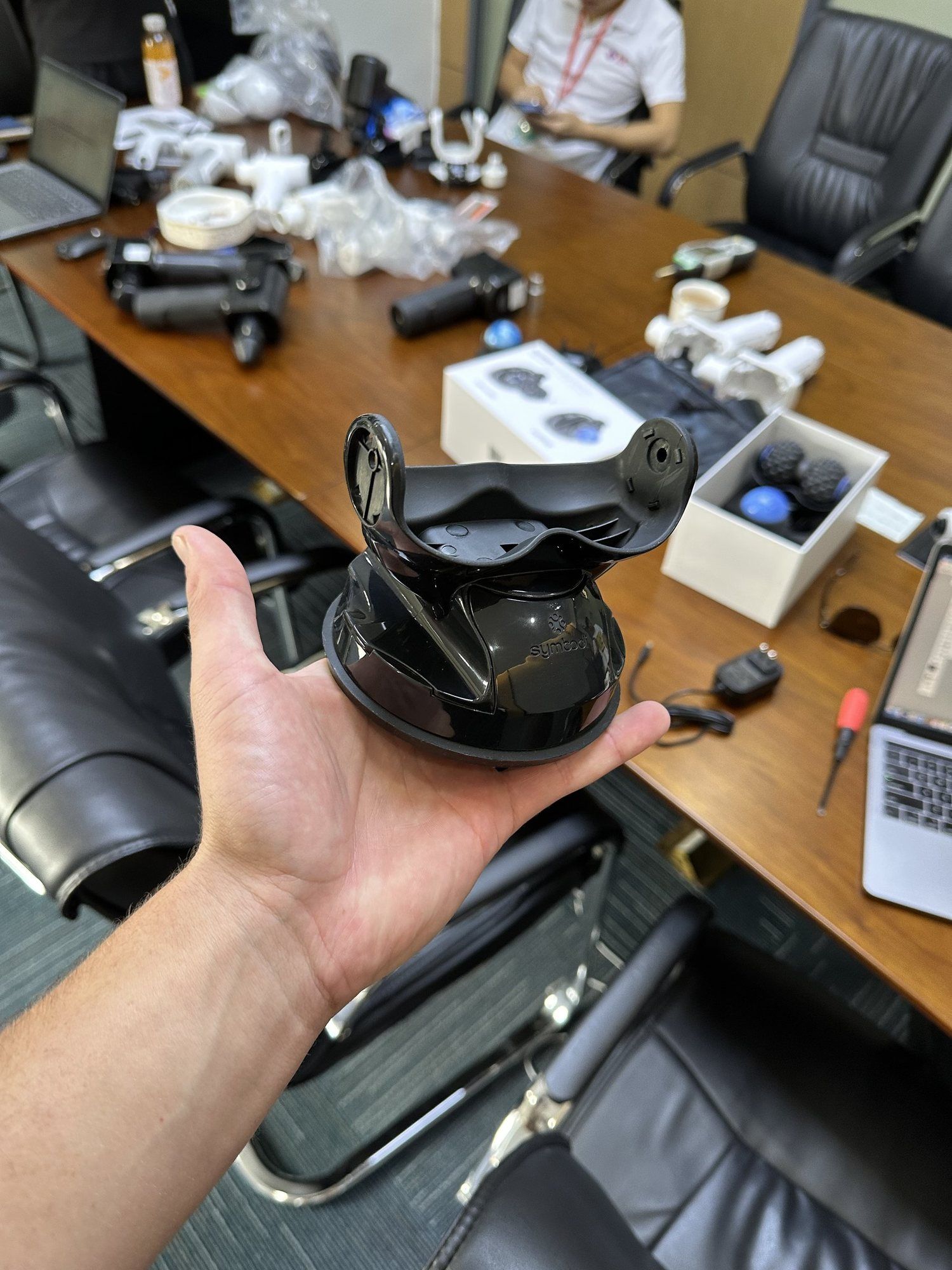 A hand holding a sample of the Vertiball wall mount over a table of parts.